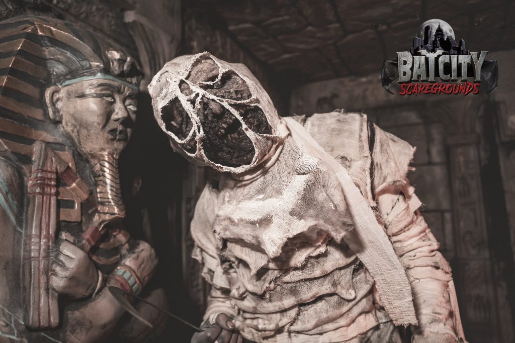 A reanimated mummy standing infront of a sacrophagus with the Bat City Scaregrounds logo on top right.
