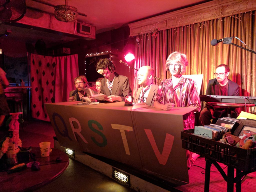Mo Mer Mo performers in funky outfits sitting behind a cardboard table imitating TV presenters.
